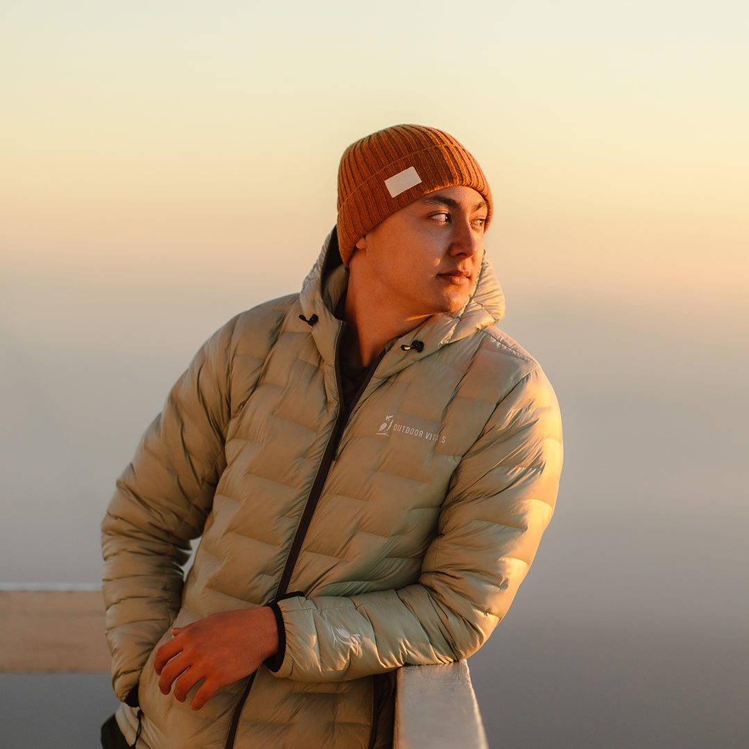 A man wearing a NovaPro down jacket and a beanie looking out at a sunset.