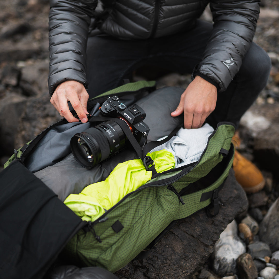 Top-down view of a person packing camera equipment, clothing, and gear inside the main compartment of the Outdoor Vitals KotaUL Ultralight Travel & Adventure Backpack while outdoors.
